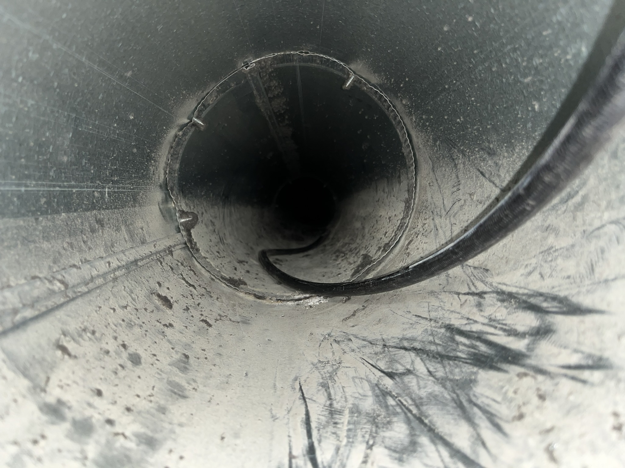 Close-up view inside a dusty metal pipe with a black cable or hose running through the center toward the dark opening.