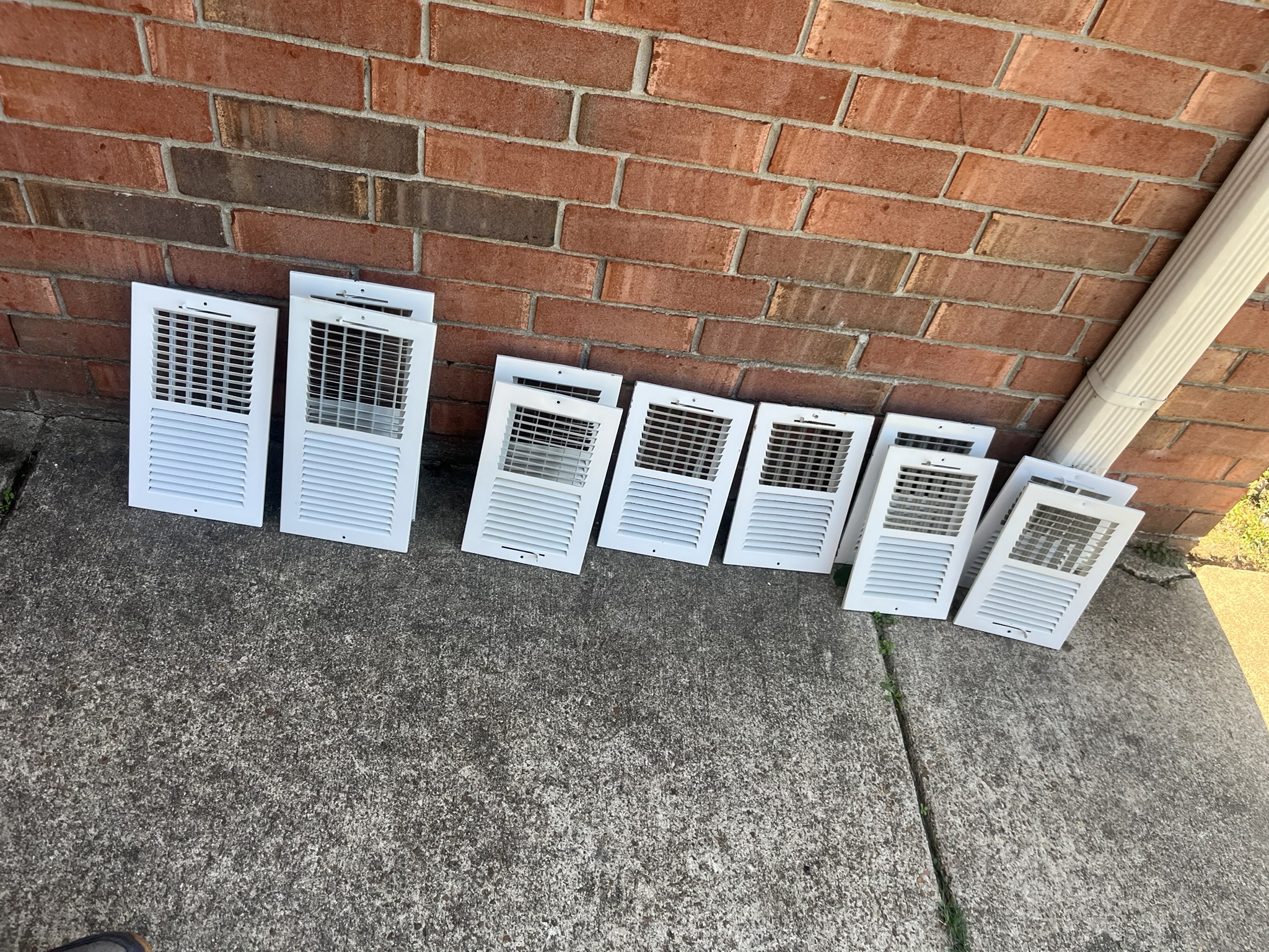 Seven white air vent covers of varying sizes are lined up against a red brick wall on a concrete surface, next to a downspout—perfect for those considering an air duct cleaning service.