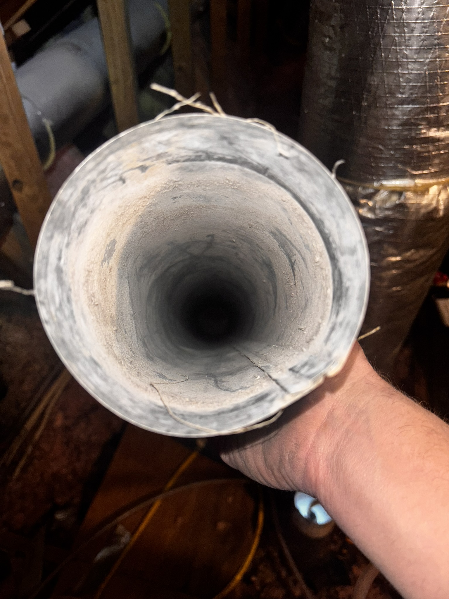 A person holds a dusty, open metal duct or pipe, viewed from the end, in what appears to be an attic or unfinished space.