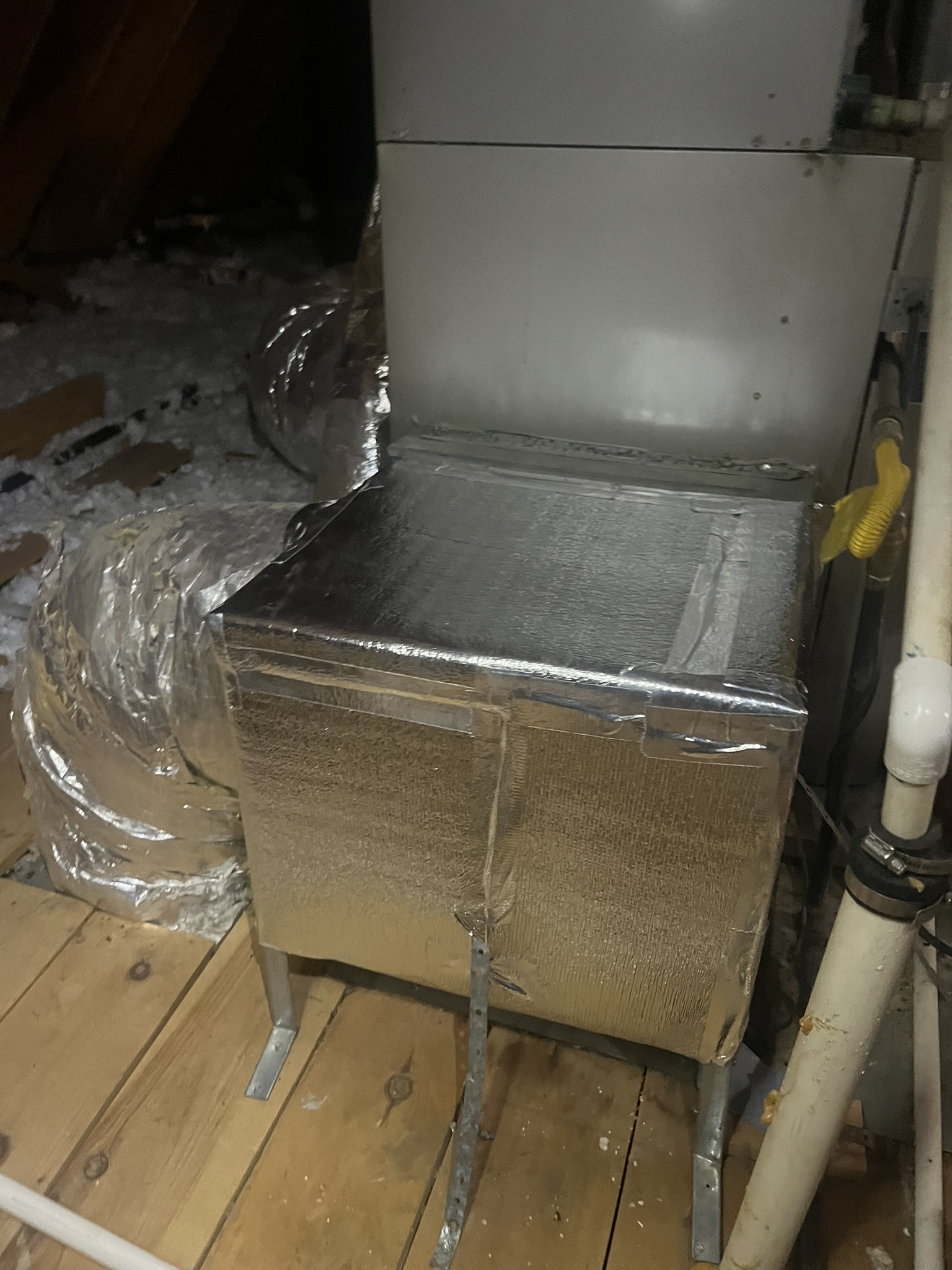 A metal HVAC unit wrapped in reflective insulation is installed on wooden beams in an attic, with ductwork and pipes nearby.